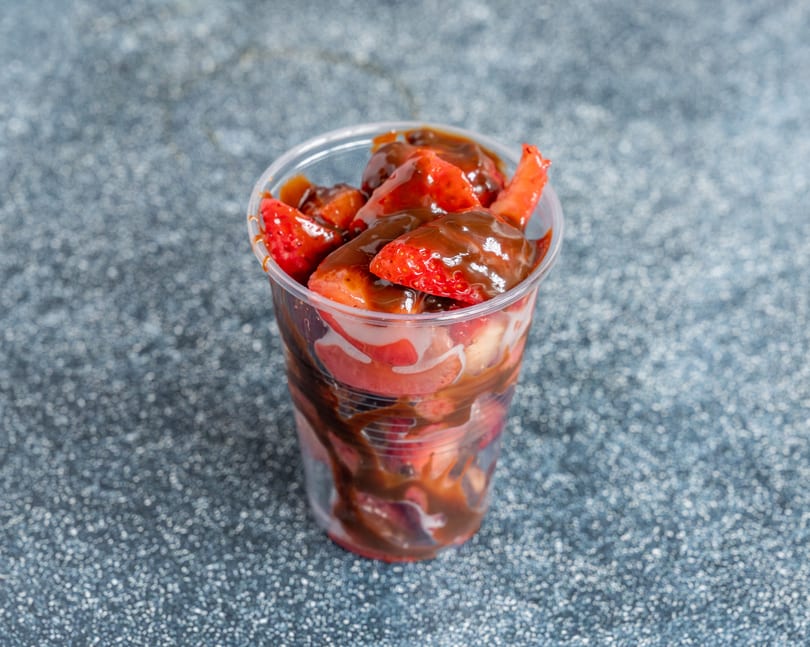 Fresh strawberries drizzled with chocolate sauce in a clear plastic cup on a speckled gray surface.