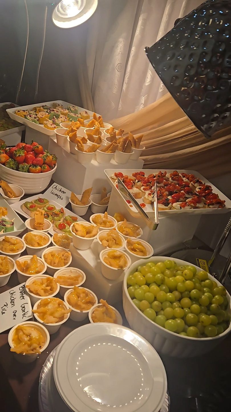 A buffet table laden with various appetizers, including bruschetta, lumpia, chicken over rice, strawberries, and grapes.